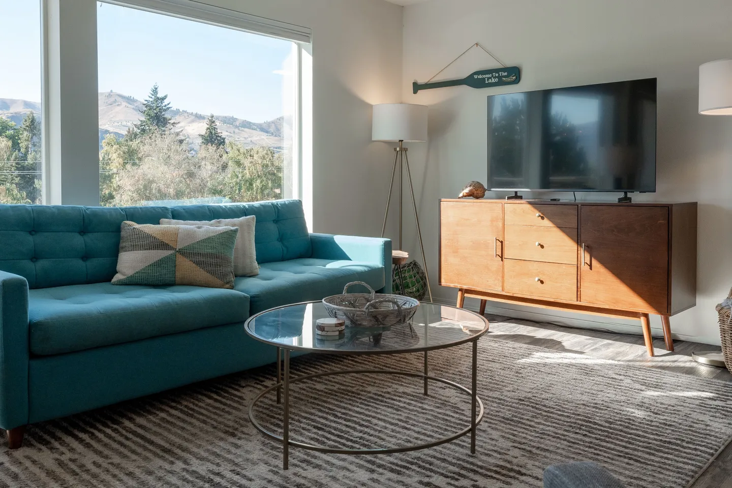 Living room featuring a turquoise sofa, a round glass coffee table, and a TV on a wooden console beside a large picture window.