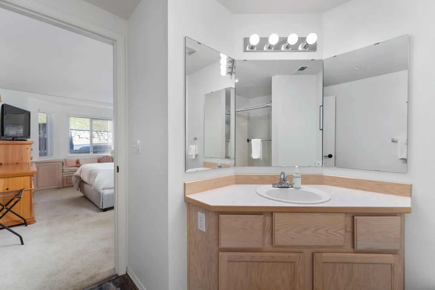 Bathroom vanity with wooden cabinets, a sink, and mirrors, with an open doorway leading to a carpeted bedroom.