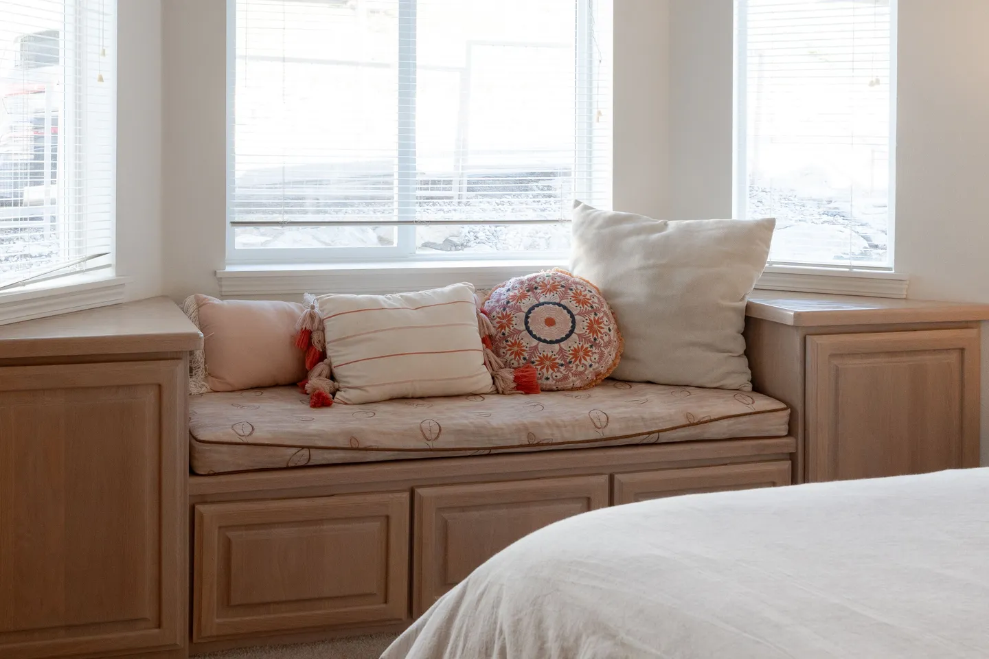 A built-in wooden bay window seat with a patterned cushion, decorative throw pillows, and lower cabinetry next to a bed.