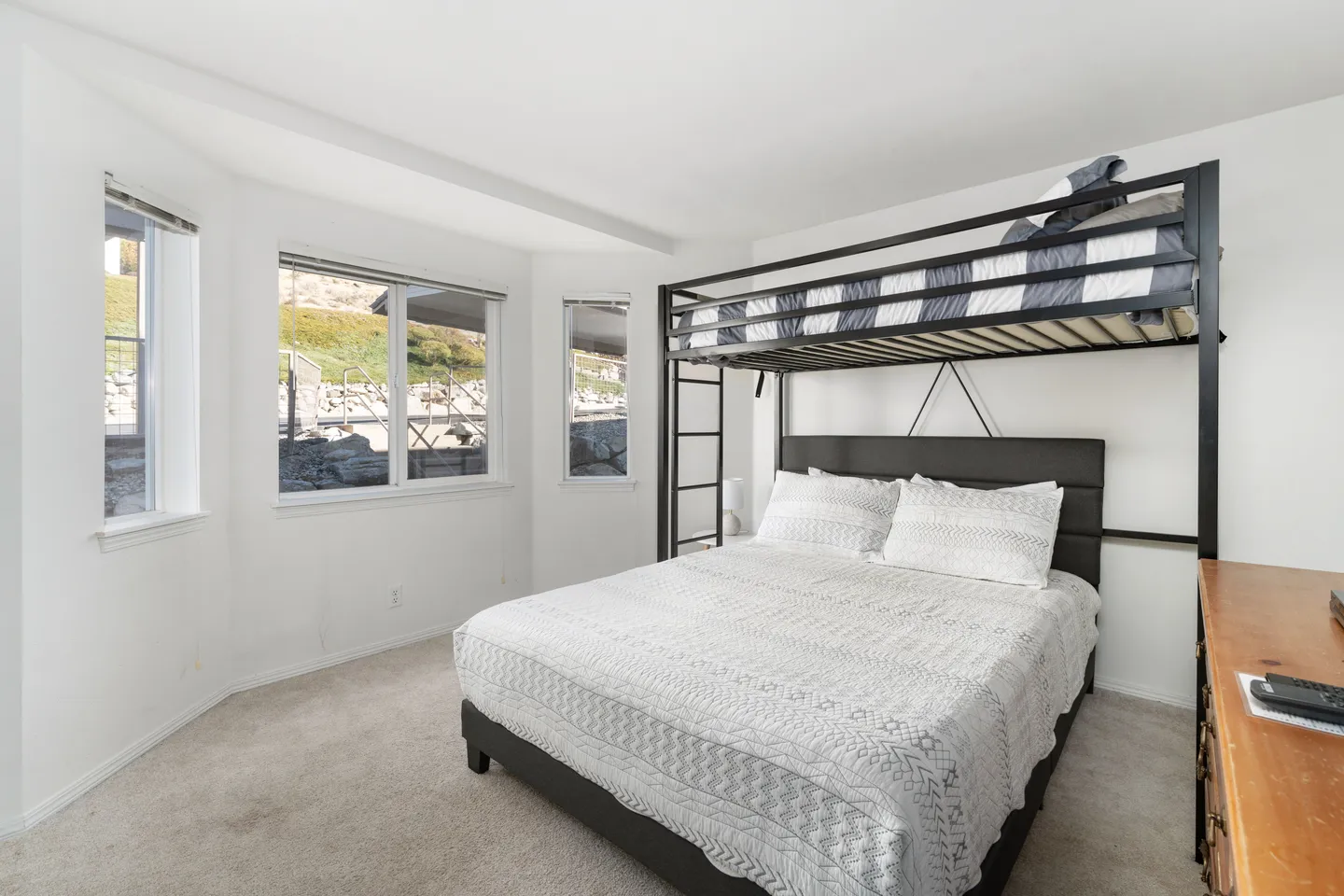 Bedroom with carpeted flooring, featuring a large bed beneath a metal loft bunk, with windows overlooking an outdoor hillside.