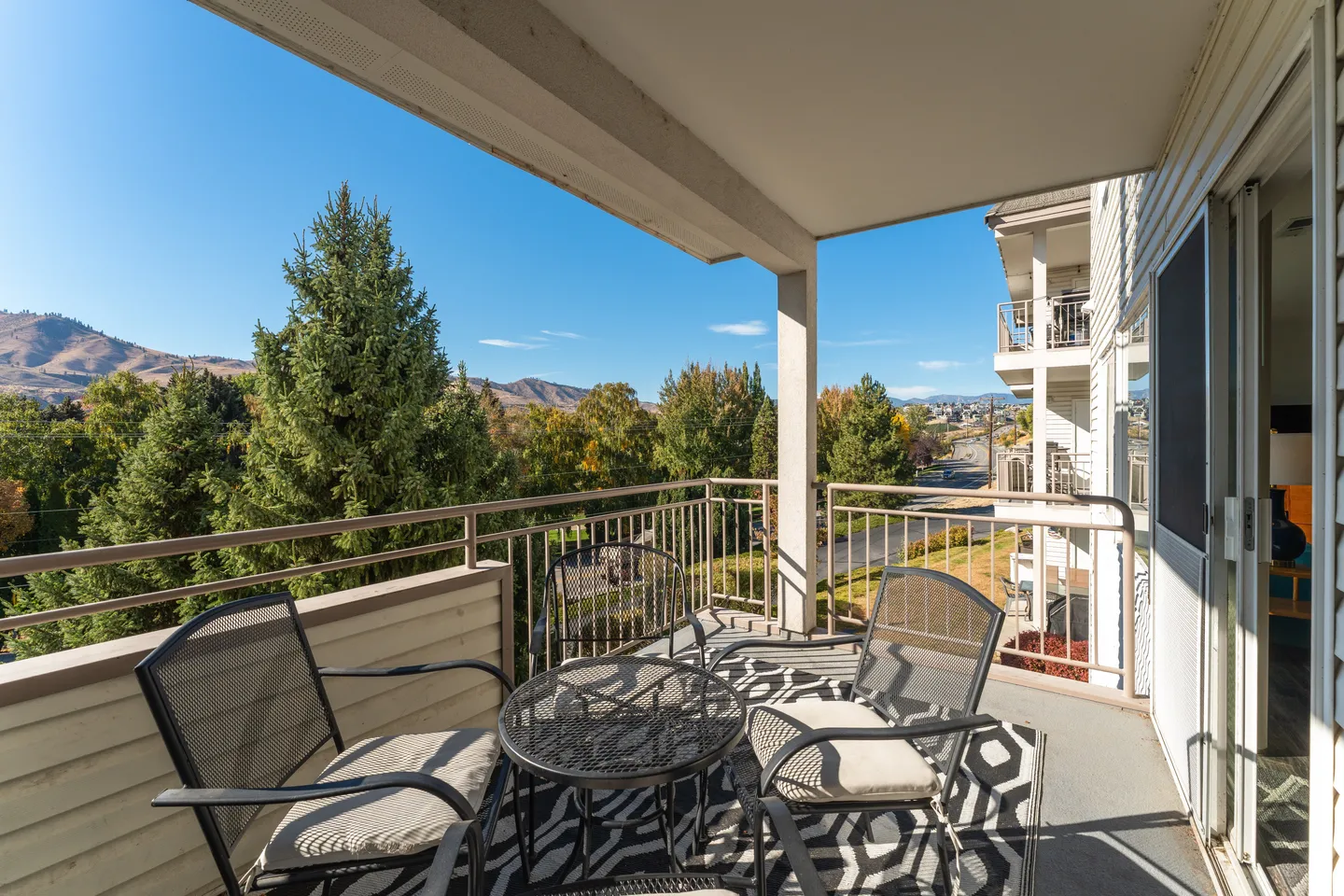 A covered balcony with an outdoor table, chairs, and geometric rug overlooking trees and rolling hills under a blue sky.