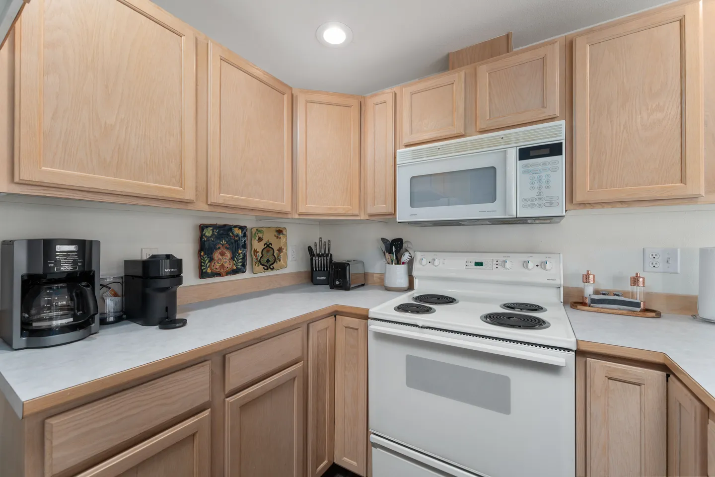Kitchen corner with light wood cabinets, white electric stove, overhead microwave, and coffee makers on the countertop.