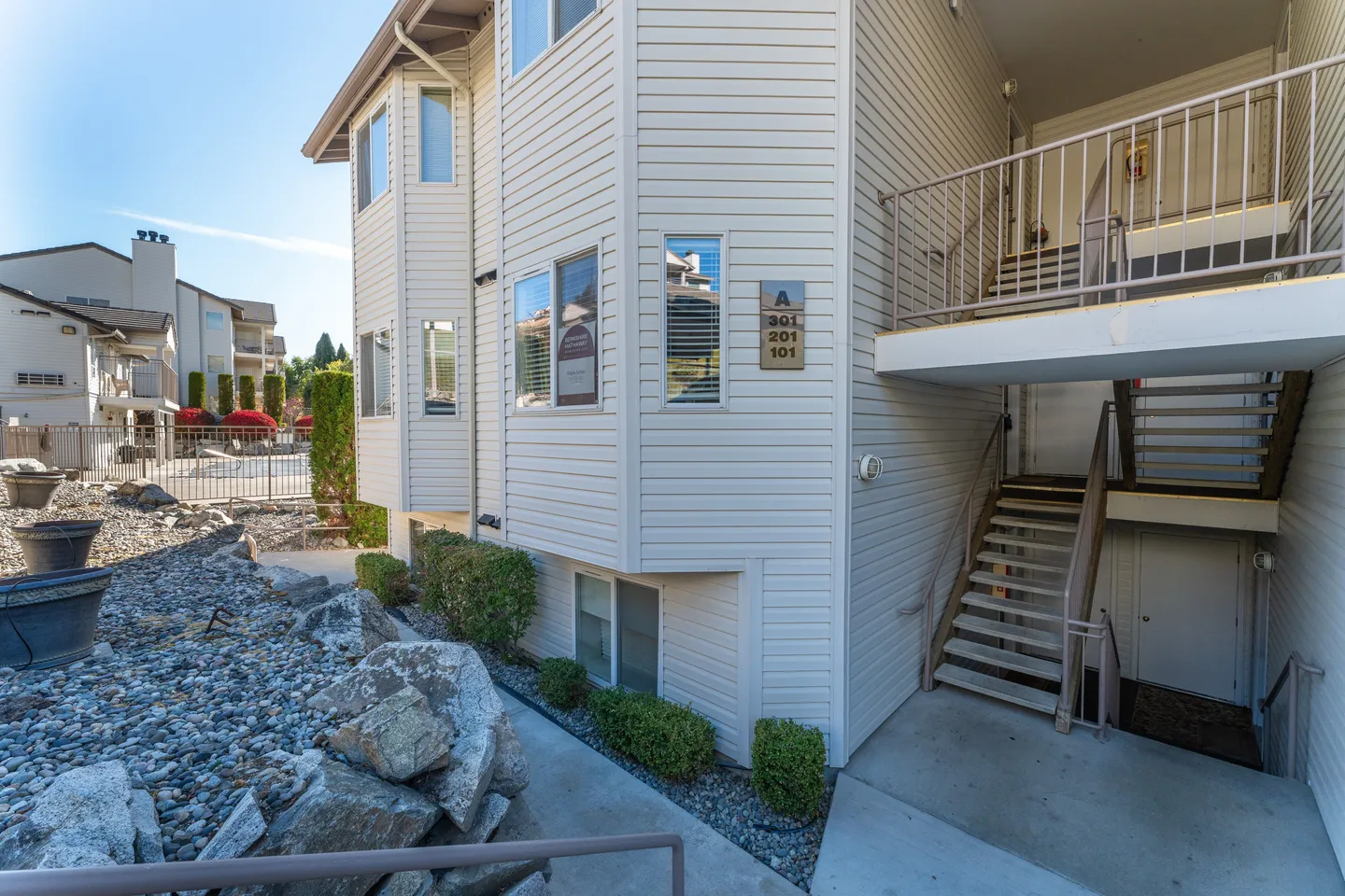 Exterior view of a multi-story residential building with light siding, an outdoor staircase, and rock landscaping.