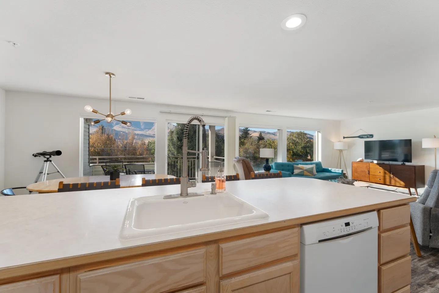 View over a kitchen island with a white sink toward the dining area, living room, and large windows.