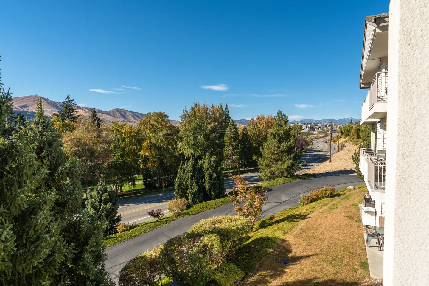 Elevated view from a building exterior overlooking paved roads, green and autumn-colored trees, and distant rolling hills under a blue sky.