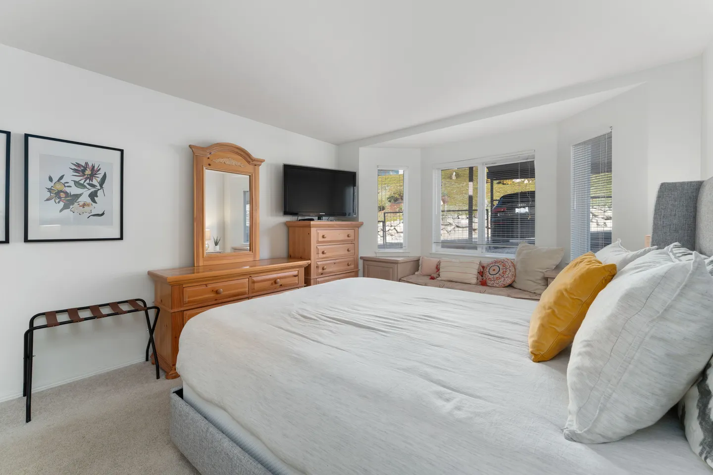 Bedroom featuring a large bed with a light-colored duvet, a wooden dresser with a mirror, a mounted TV, and a bay window bench.
