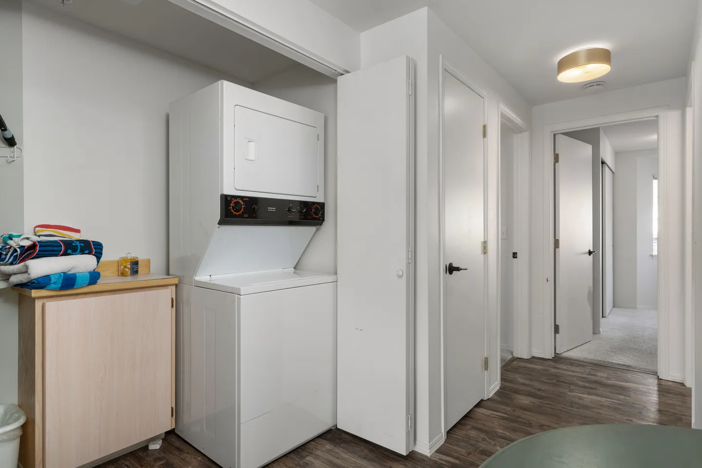 A stacked white washer and dryer unit in a closet area next to a hallway with wood-look flooring.
