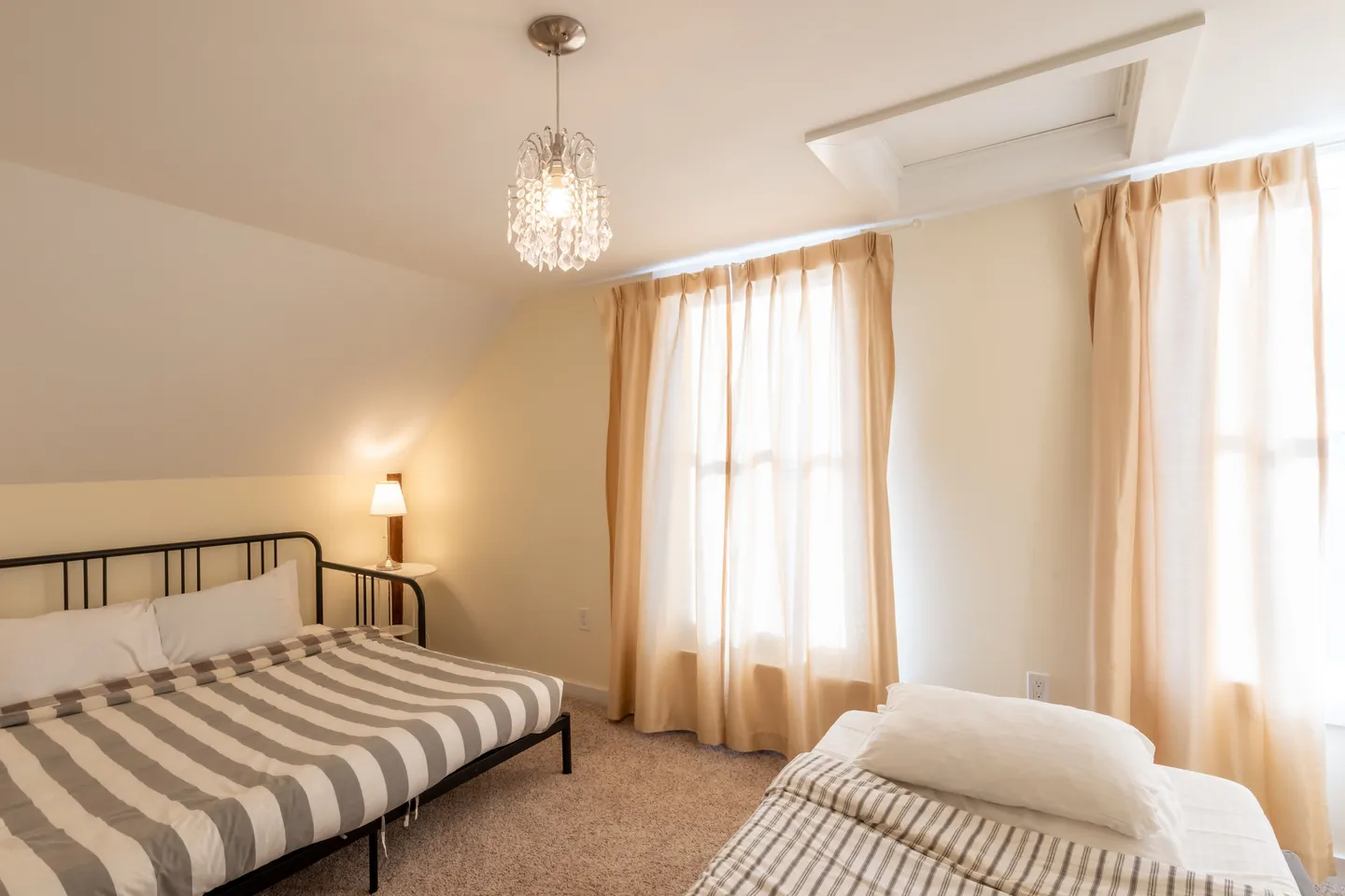 Bedroom with sloped ceiling, two beds with striped bedding, carpeted floor, and windows with sheer curtains.