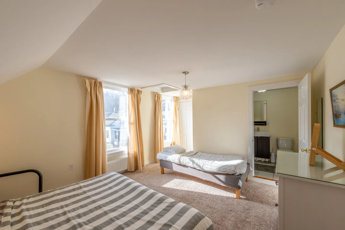 Bedroom with carpeted flooring, two beds with striped bedding, windows, and an en-suite bathroom visible through an open doorway.