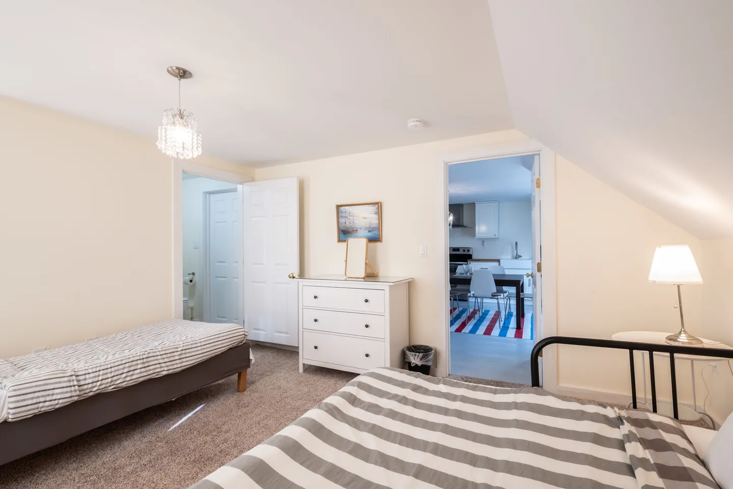 Bedroom with carpet, two beds, a white dresser, and open doors leading to a bathroom and a dining area.