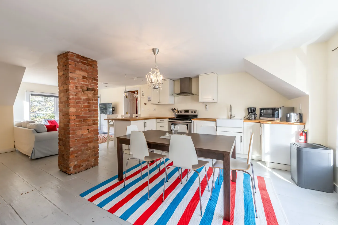 Open-concept dining and kitchen area featuring an exposed brick pillar, dining table with modern chairs over a striped rug, and white kitchen cabinetry.