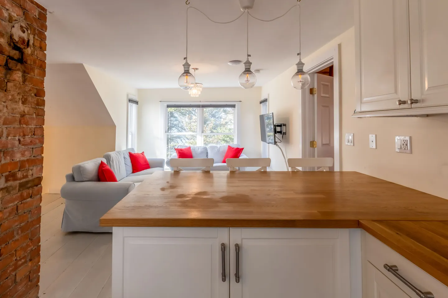 Open-concept kitchen island with wooden countertop looking into a living room with couches, red pillows, and an exposed brick column.