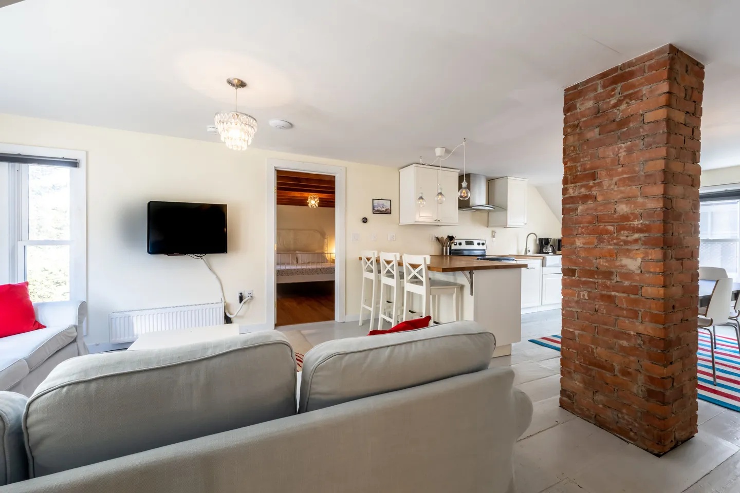 Open-concept living room with a gray sofa facing a kitchen peninsula with barstools, an exposed brick column, and a doorway leading into a bedroom.