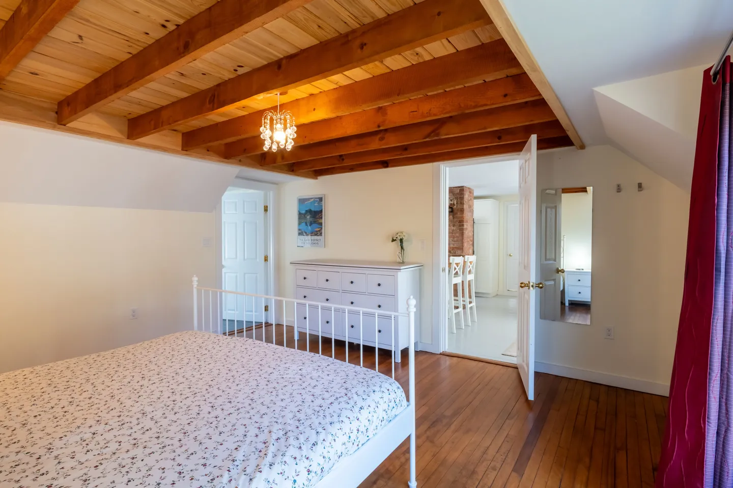 Bedroom with wood-beamed ceiling, wood floor, white metal frame bed, and view through open door to kitchen.