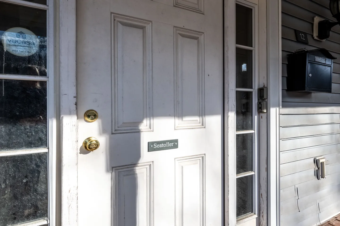 Close-up of a white paneled front door with a brass doorknob, a plaque reading 'Seatoller', sidelight windows, and a black mailbox on the adjacent siding.