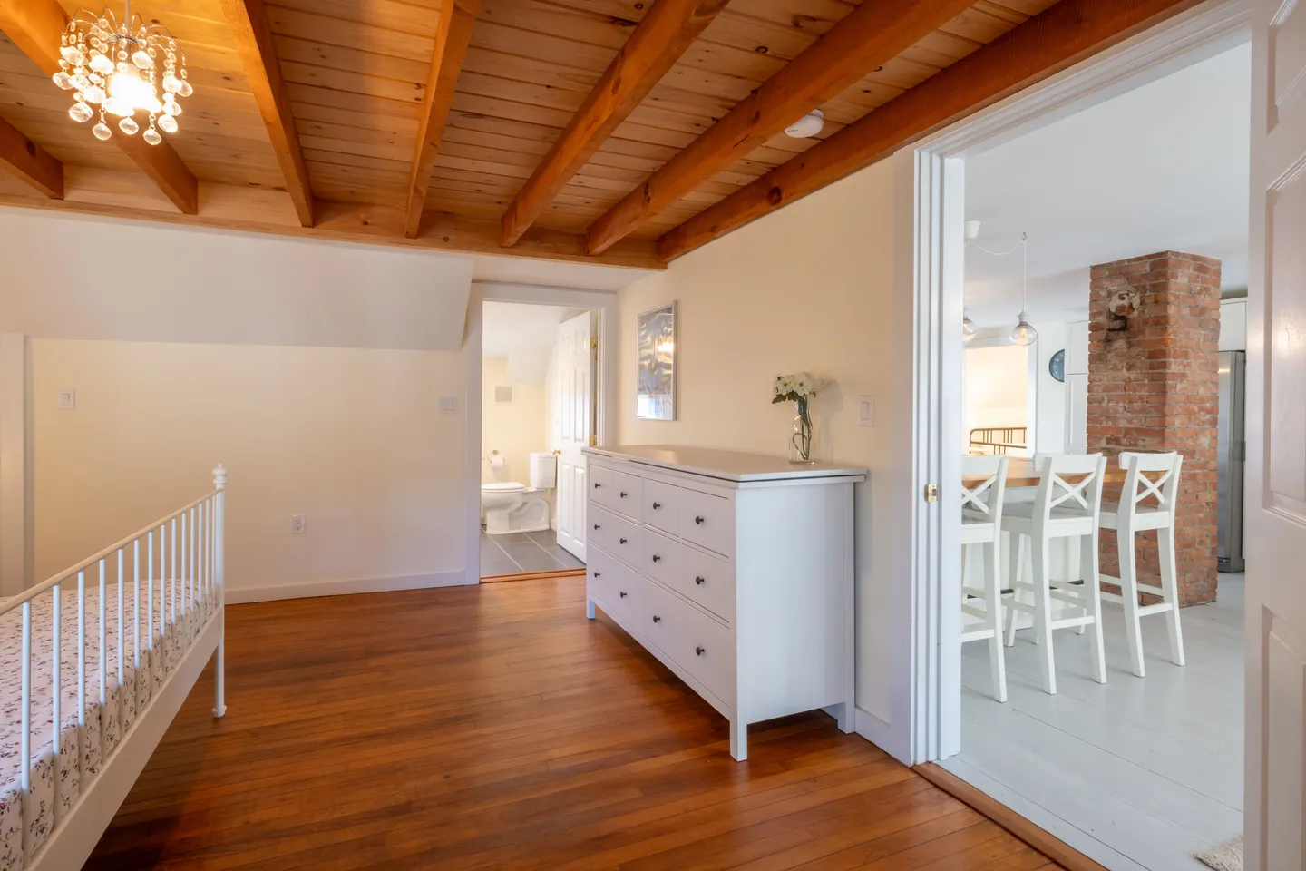 Bedroom with hardwood floors and exposed wood beam ceiling, showing a white dresser, bed frame, an open doorway to a bathroom, and another doorway to the kitchen.