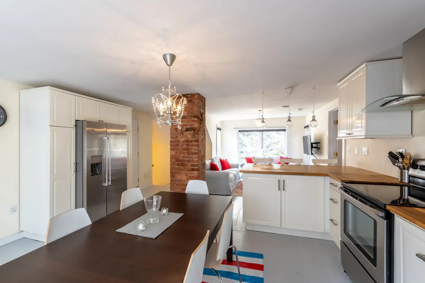 Open-concept dining area and kitchen with a dark wood table, stainless steel appliances, and view of the living room beyond an exposed brick column.