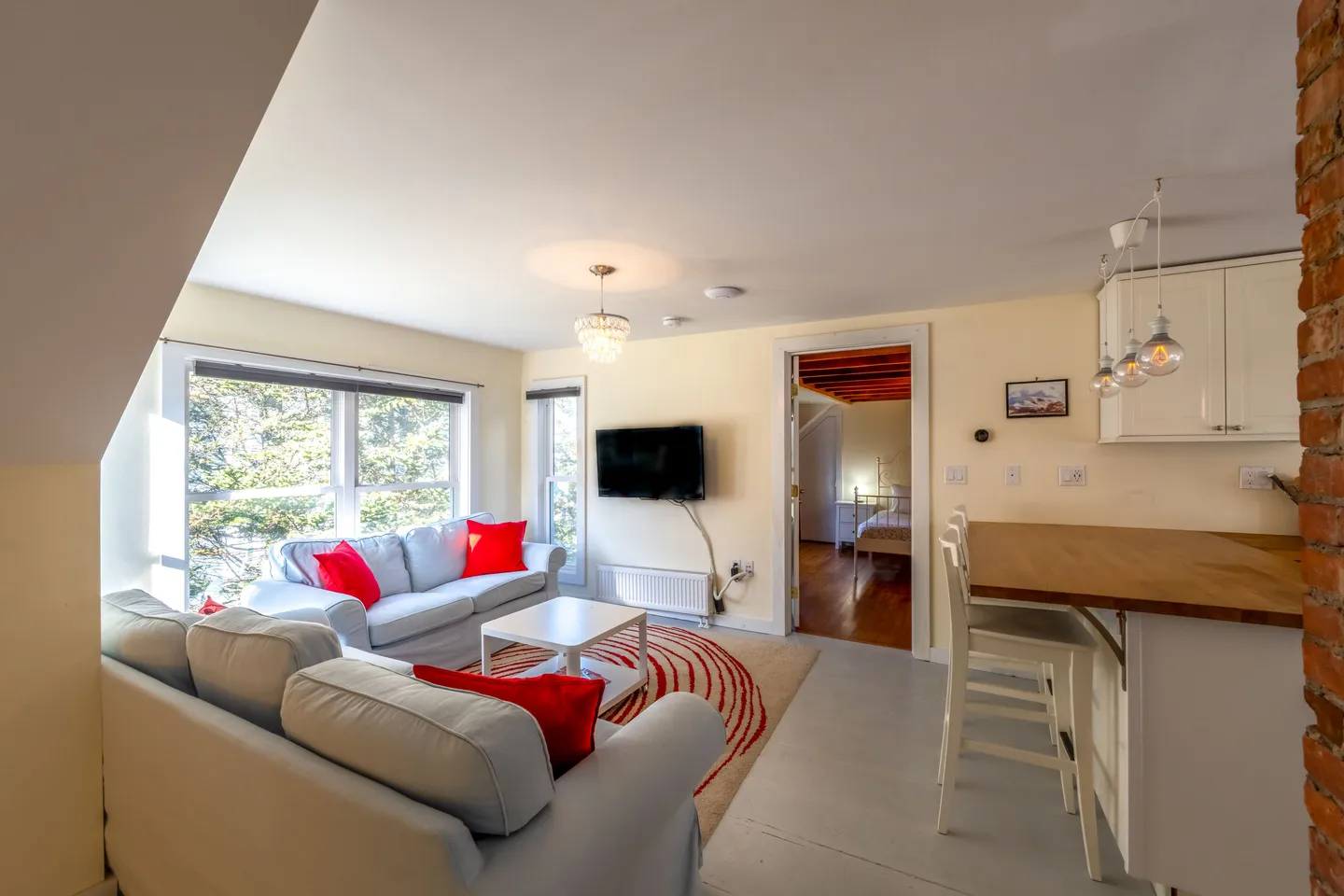 Open living area with two gray sofas accented by red pillows, a coffee table, a wall-mounted TV, and an adjacent kitchen breakfast bar.