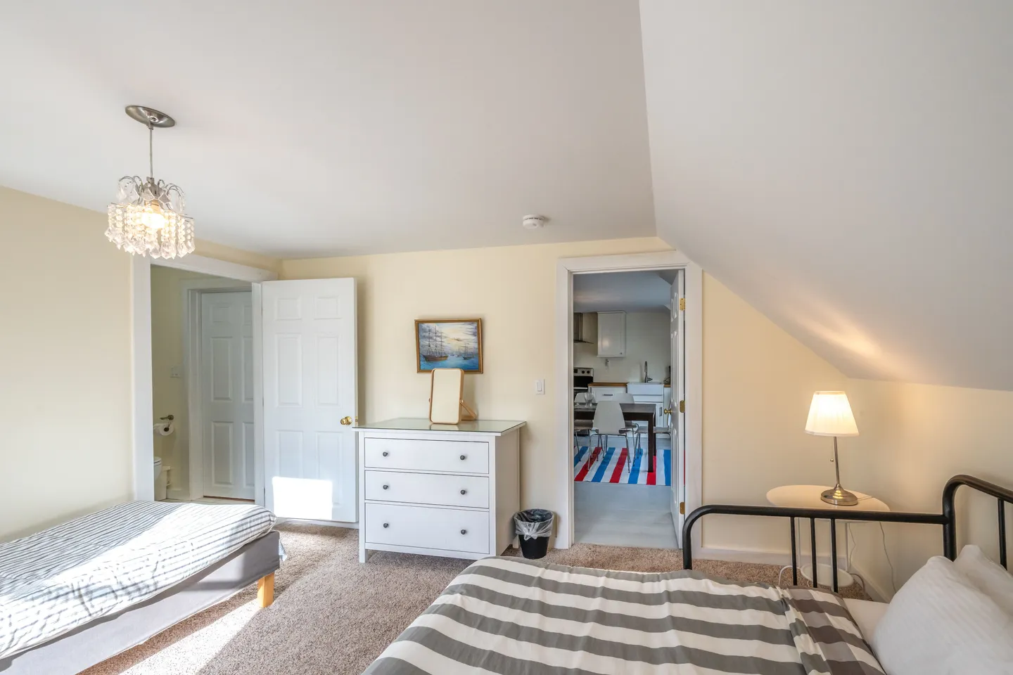 Bedroom featuring two beds, carpeted floor, a white dresser, and an open doorway looking into a kitchen and dining area.