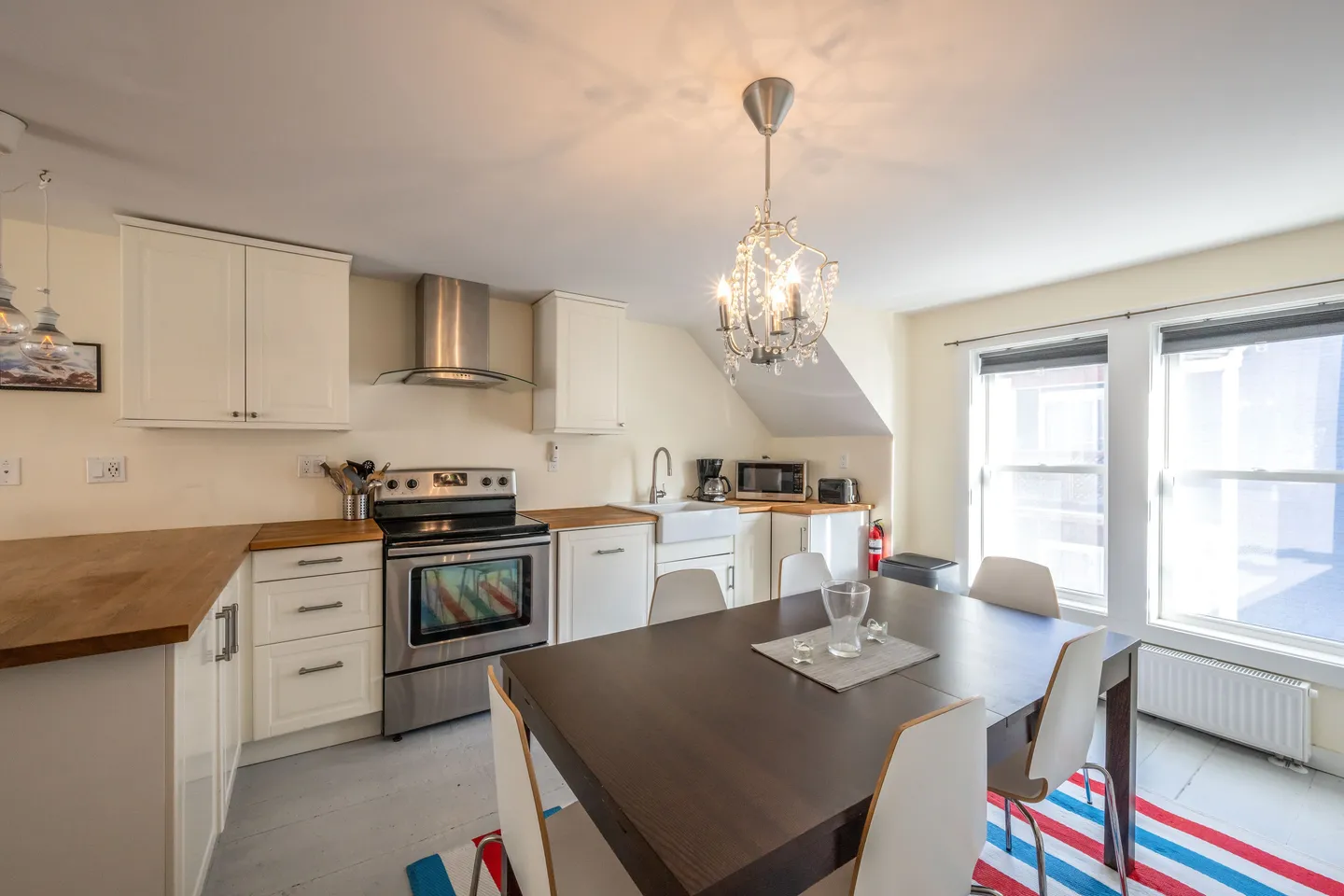 Open kitchen and dining space with white cabinets, stainless steel appliances, a chandelier over a dark dining table, and windows along the wall.