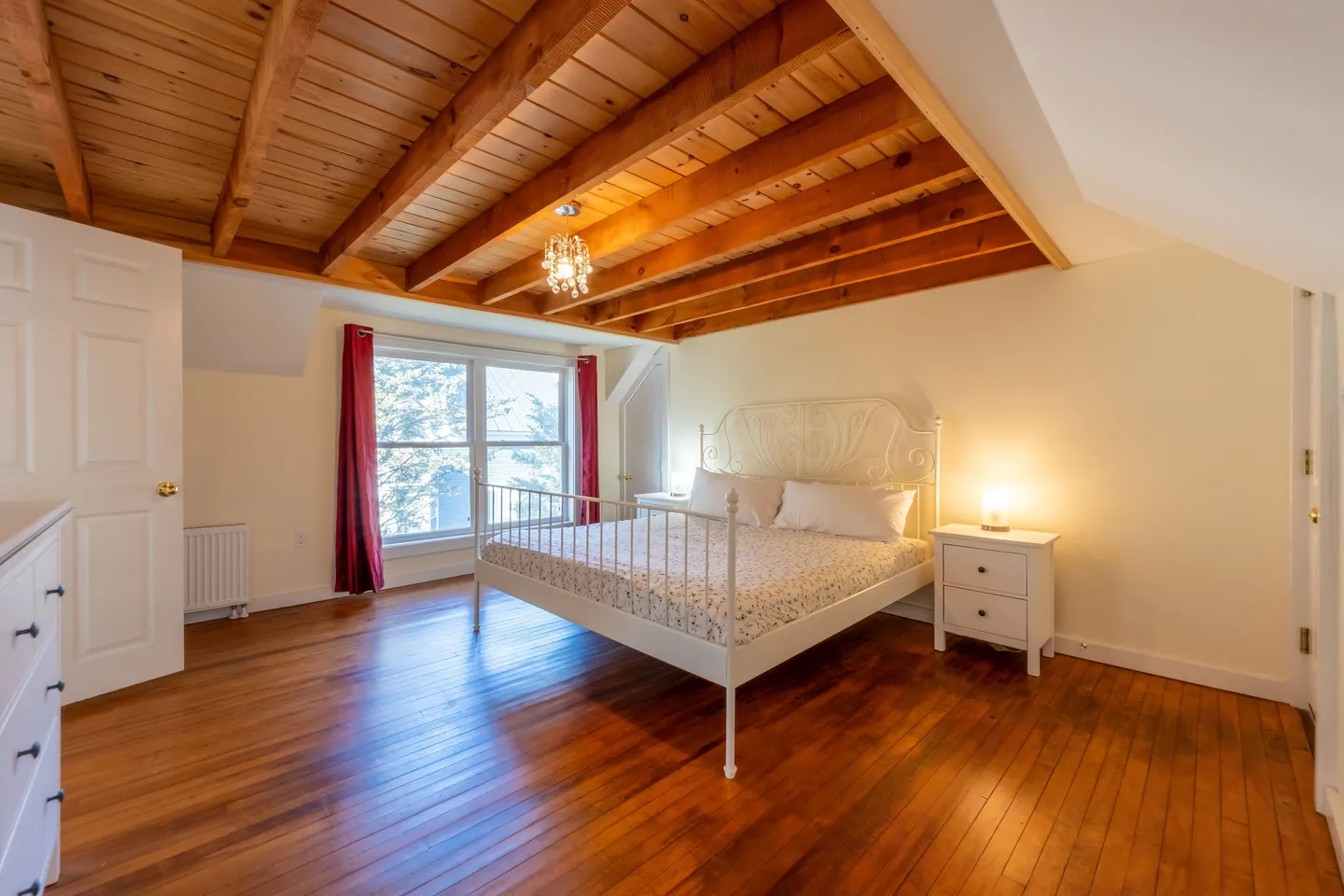 Bedroom featuring an exposed wood-beam ceiling, wood plank floors, a white metal spindle bed, and two windows framed by red curtains.