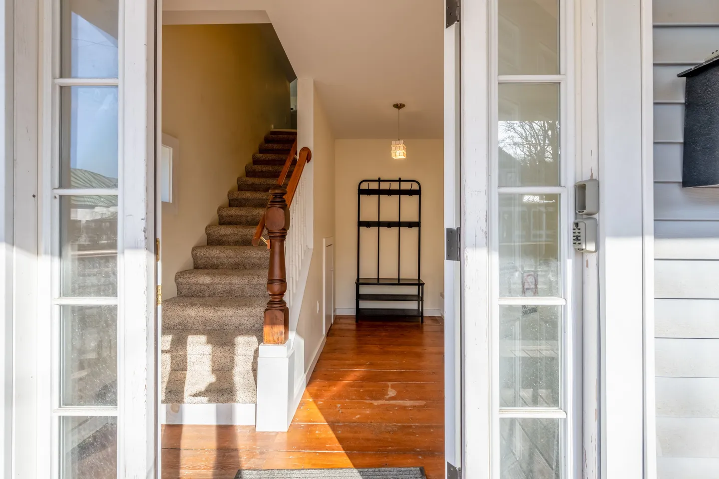 Entryway viewed through an open doorway, showing wood flooring, a carpeted staircase with a wood banister, and a black coat rack bench.