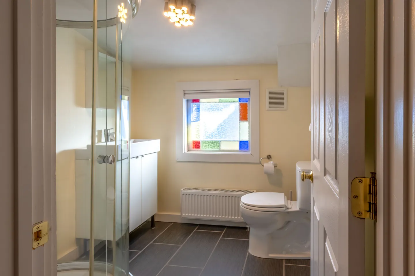 Bathroom framed by an open doorway, featuring a glass corner shower, toilet, white vanity, dark tile floor, and a window with colorful glass borders.