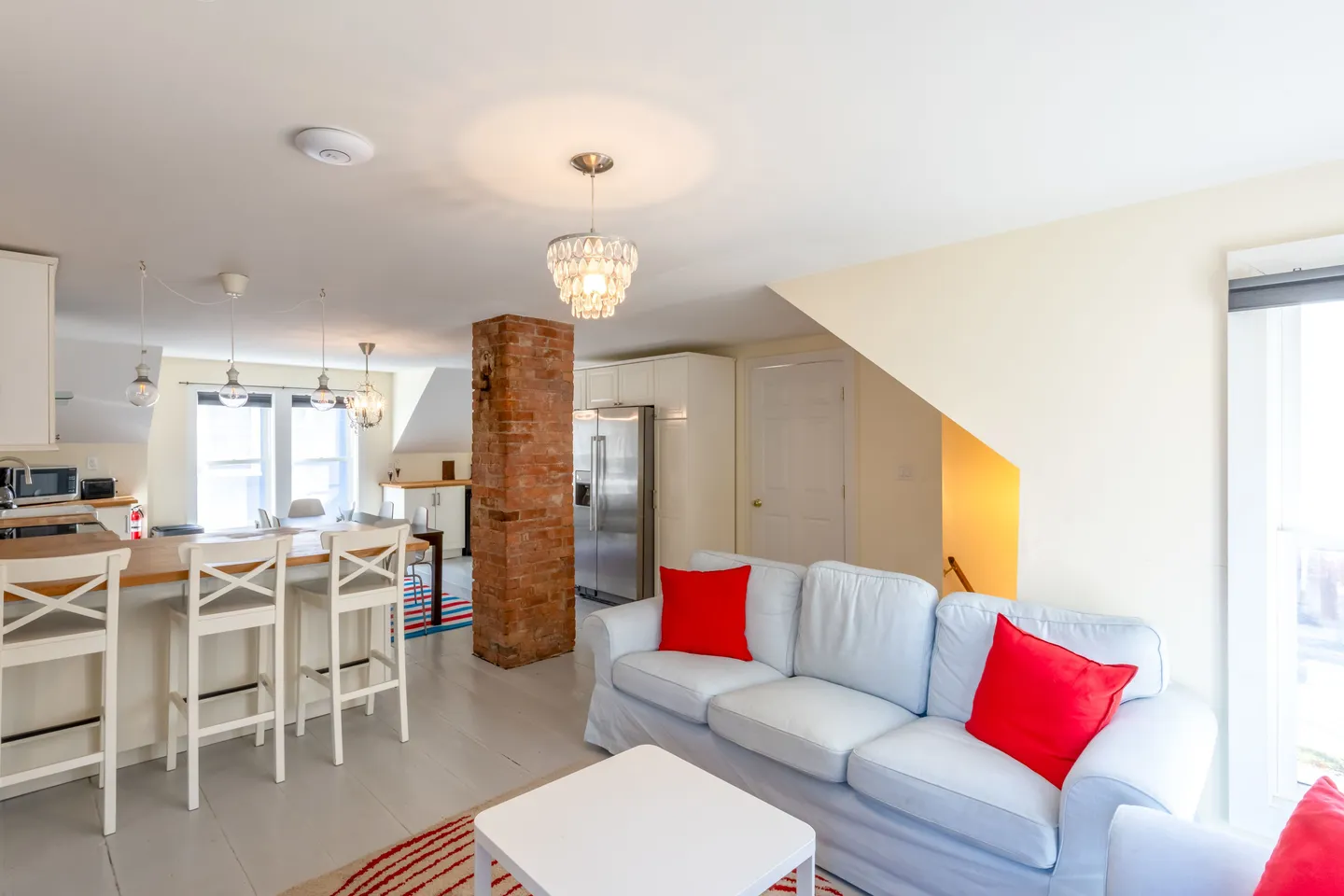 Open-concept living room and kitchen featuring a white sofa with red accent pillows, exposed brick pillar, and breakfast bar with stools.