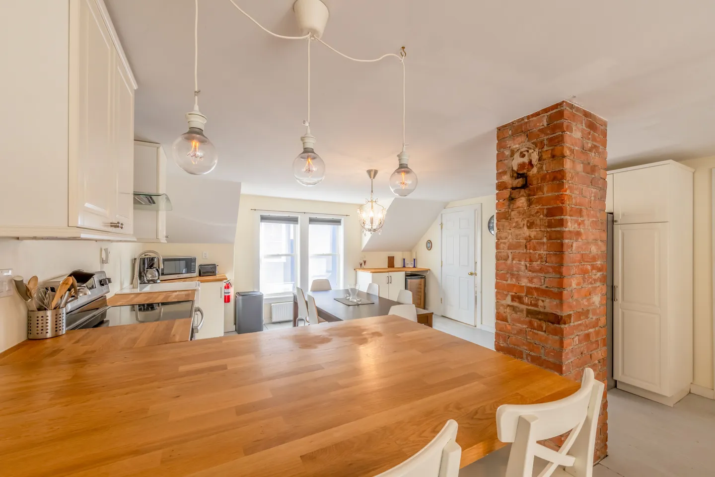 Open-concept kitchen and dining area featuring a wooden breakfast bar, hanging pendant lights, and an exposed brick pillar.