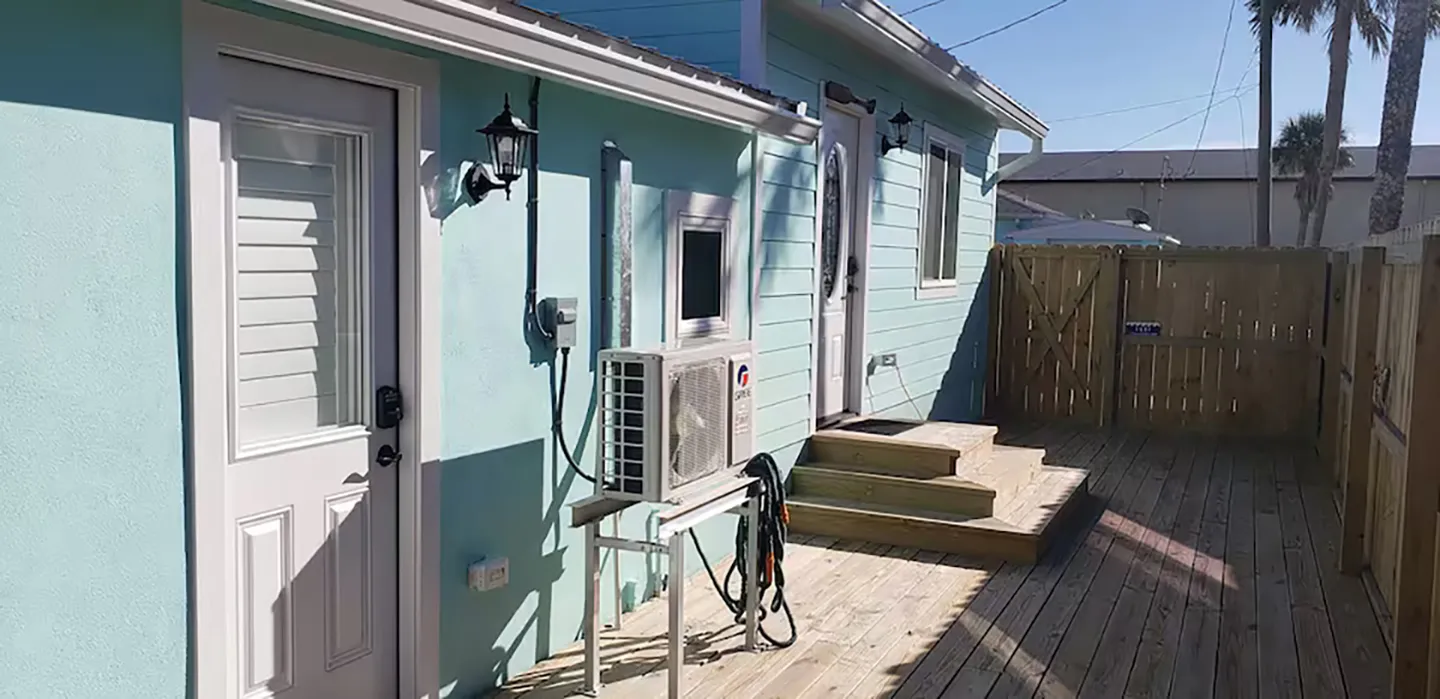 Exterior of a light turquoise home featuring a wooden deck, privacy fence, two entry doors, and a mounted air conditioning unit.