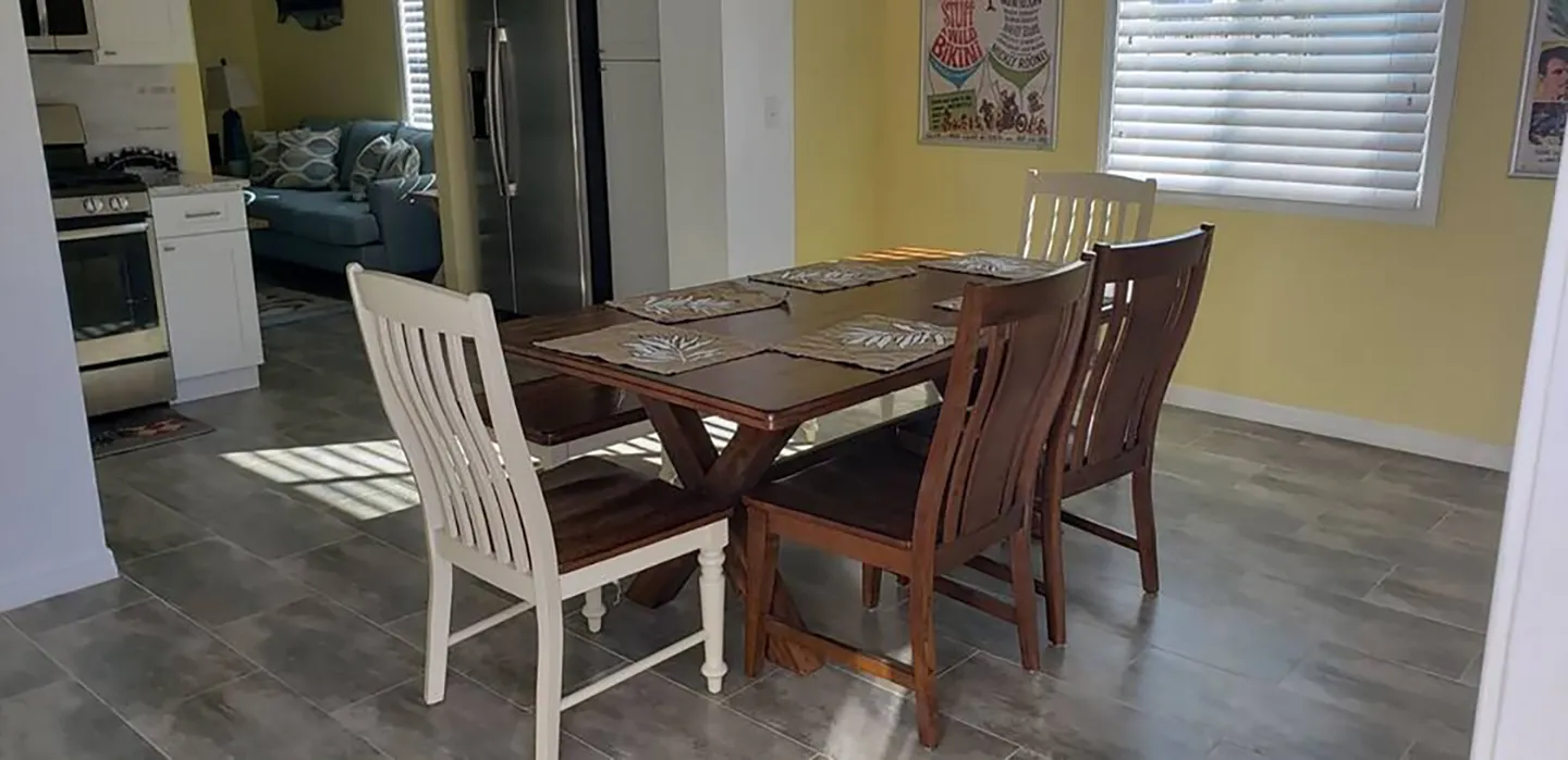 Dining area with a wooden table, mixed wooden and white slat-back chairs, and placemats, set on a tiled floor next to pale yellow walls.