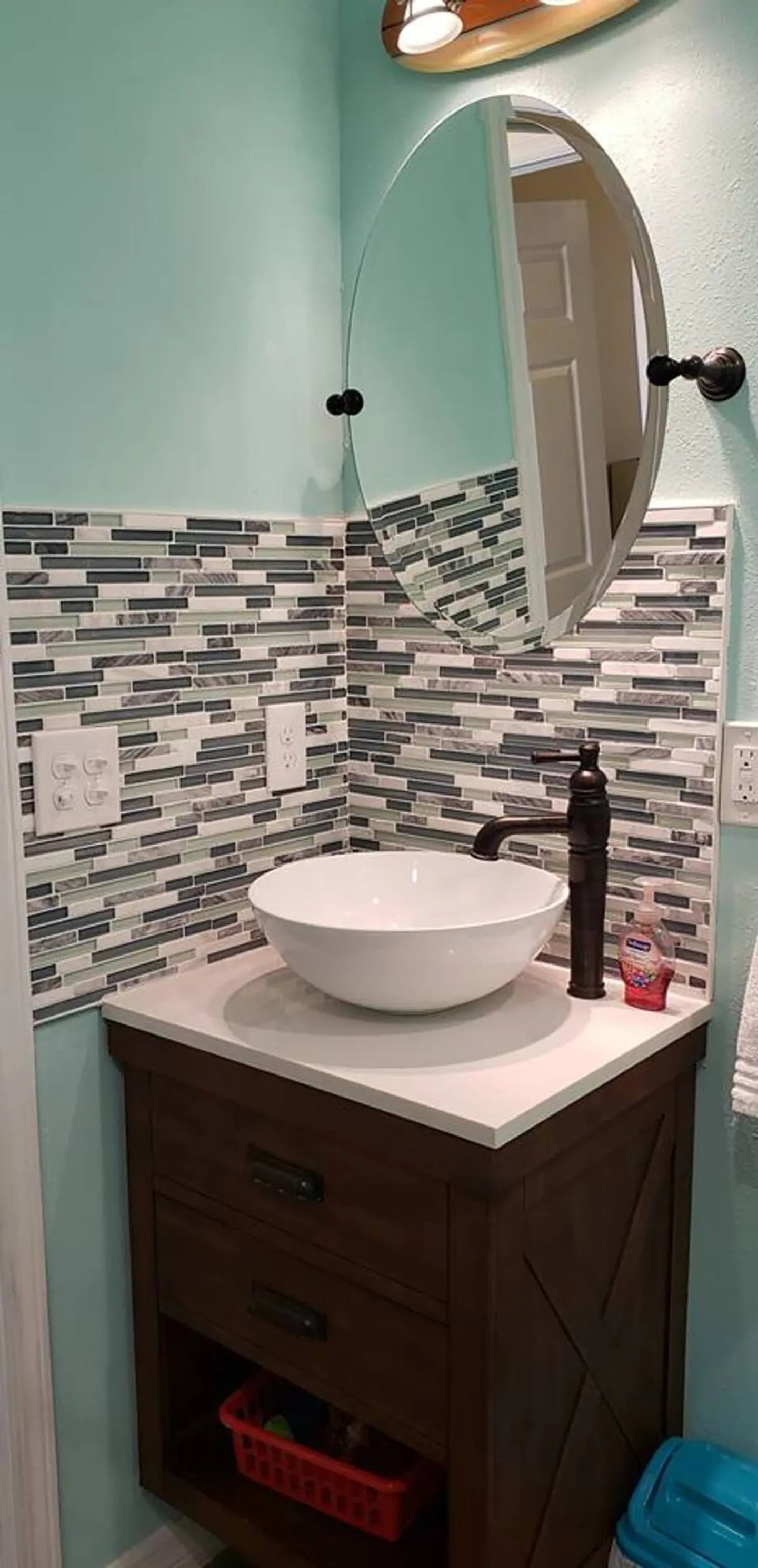 Bathroom vanity with a white vessel sink, dark faucet, oval tilting mirror, and mosaic tile backsplash against light blue-green walls.