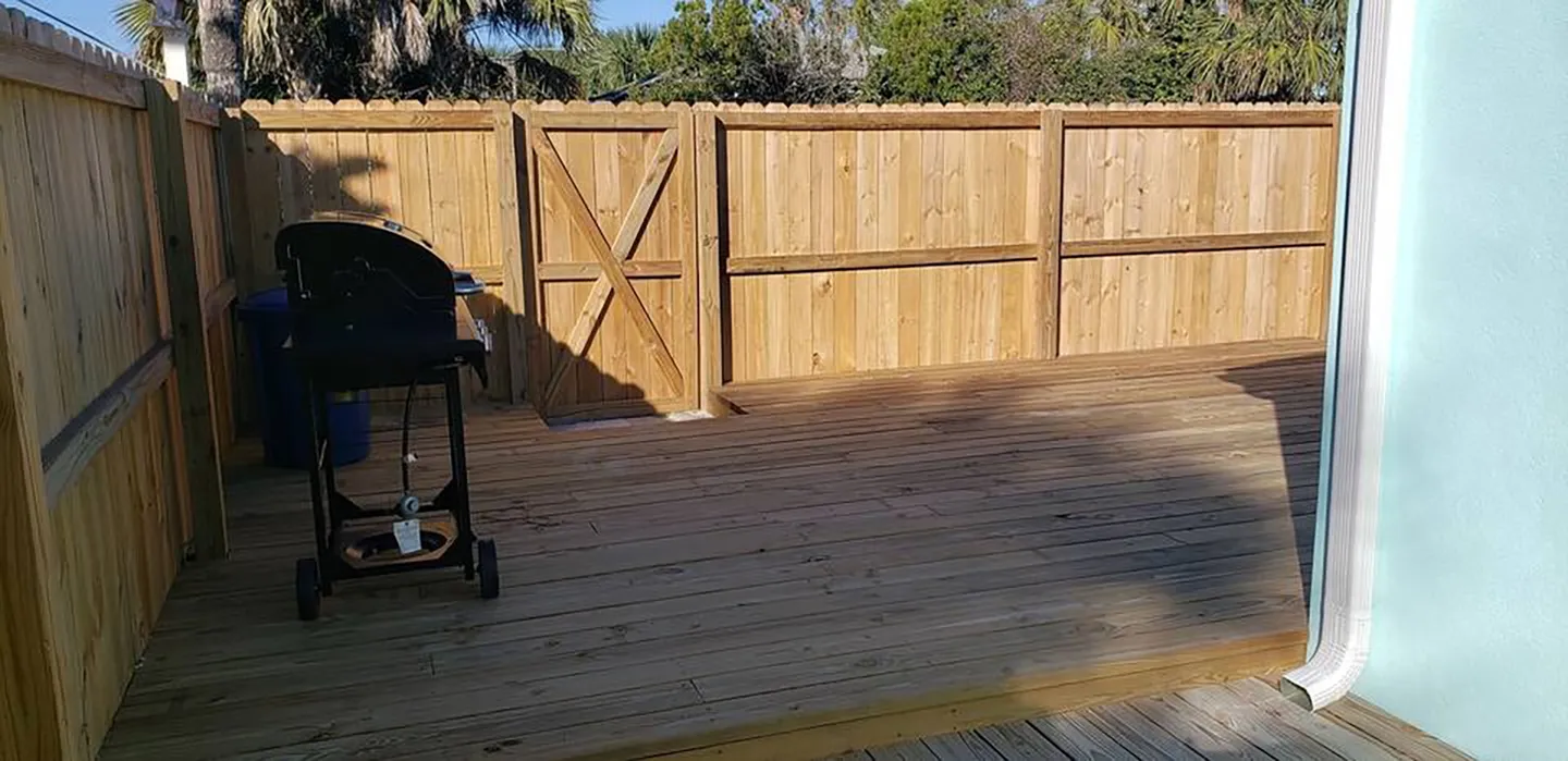 Fenced-in wooden deck with a freestanding barbecue grill and a wooden privacy gate under a clear sky.