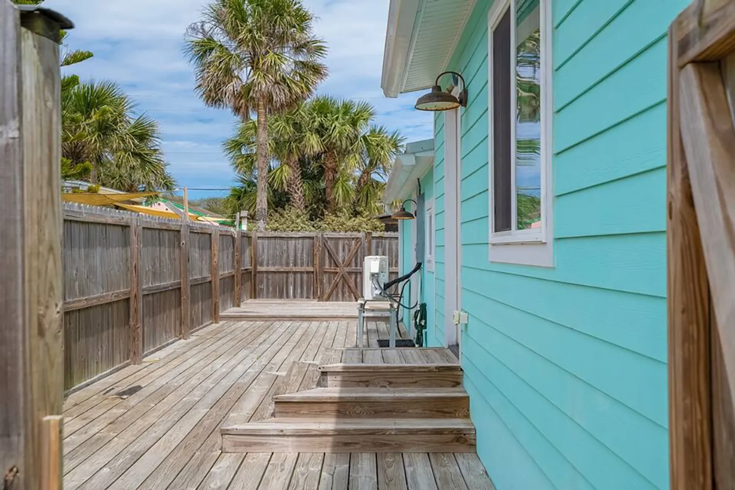 A long wooden deck alongside a turquoise exterior wall enclosed by a tall wooden fence, with palm trees visible under a blue sky.