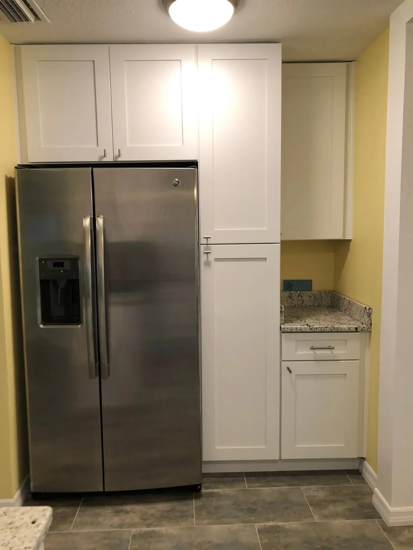 Kitchen nook featuring a stainless steel refrigerator flanked by white shaker cabinets and a small granite countertop section.