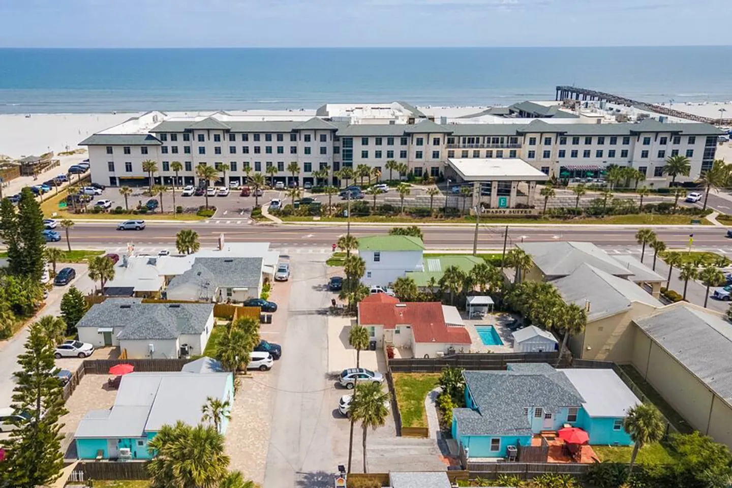 Aerial view of a coastal neighborhood with houses, a multi-story hotel across the street, a sandy beach, and the ocean.