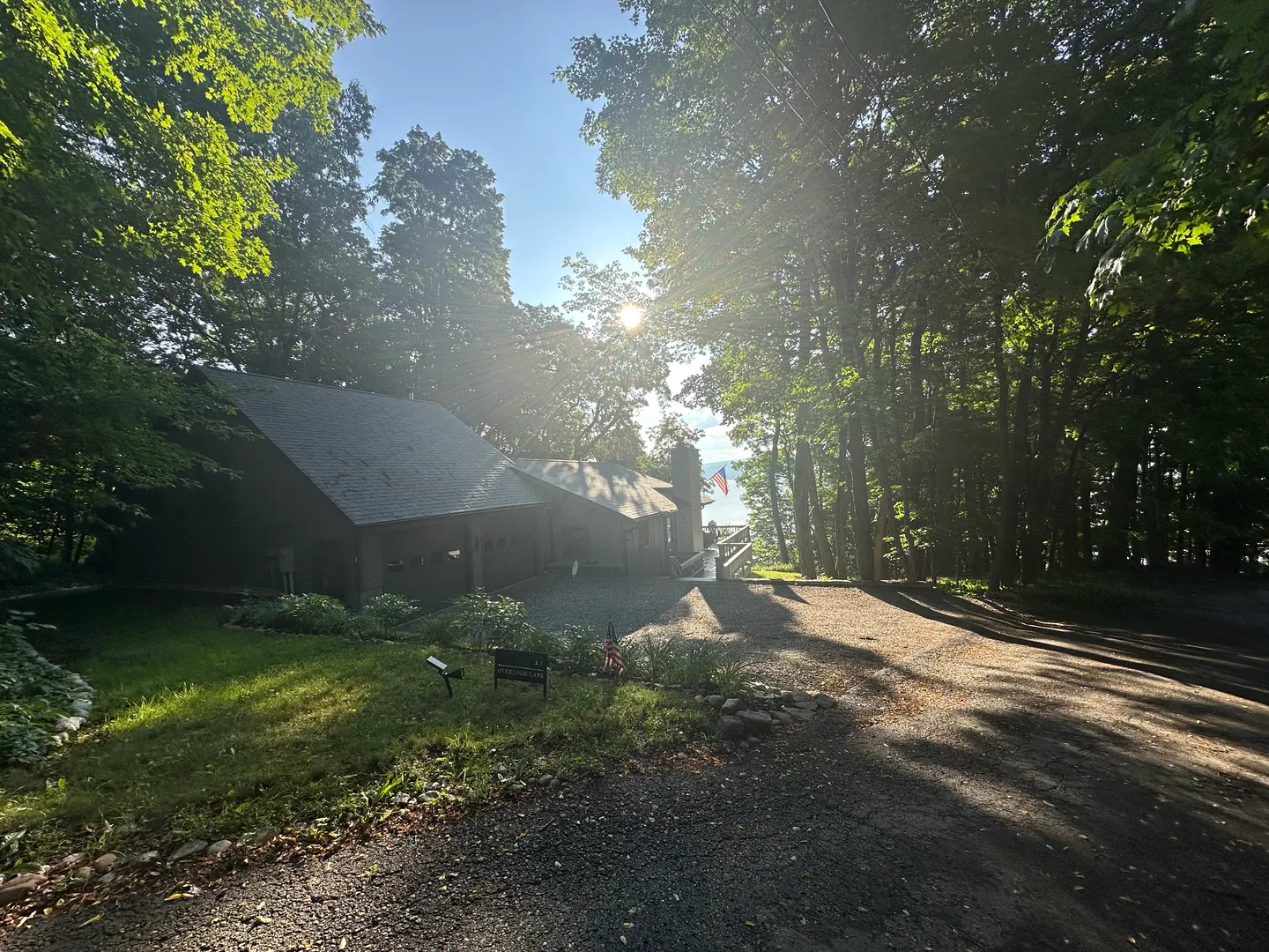 Exterior view of a dark-sided home nestled in mature trees with a gravel driveway and sunlight filtering through the canopy.