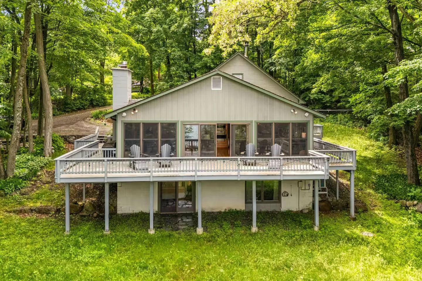 Exterior view of the back of a multi-level home featuring an elevated wooden deck with Adirondack chairs, surrounded by dense green trees.