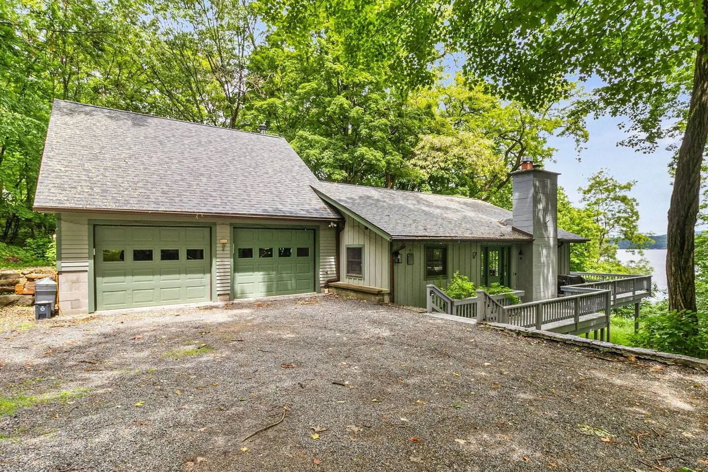 Exterior of a house with green garage doors, gravel driveway, and a wooden deck overlooking water surrounded by lush trees.
