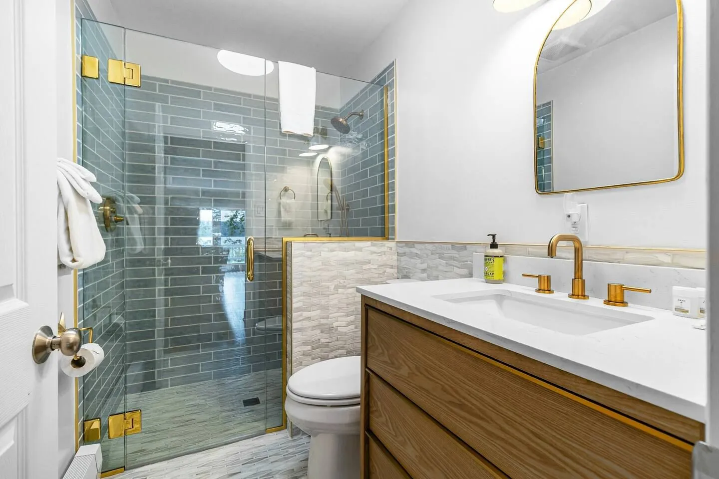 Bathroom featuring a walk-in glass shower with blue subway tile, a wooden vanity with a white countertop and gold fixtures, and an arched mirror.
