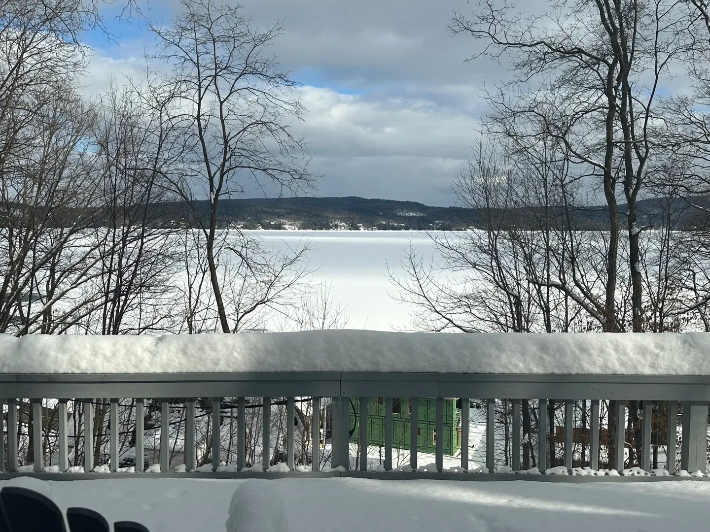 Snow-covered outdoor deck railing overlooking a frozen lake and distant wooded hills under a cloudy sky.