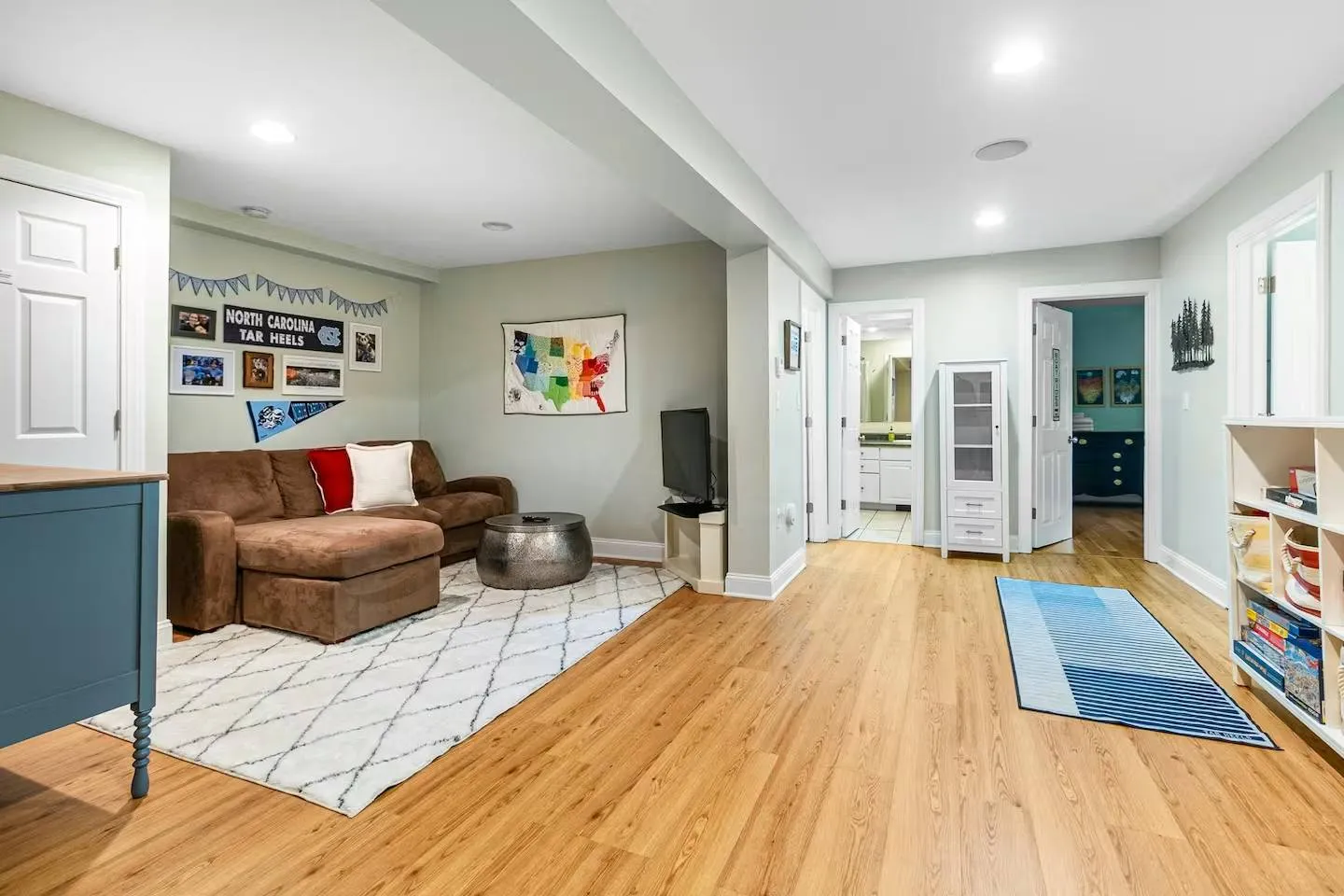 Finished basement living area with wood-look floors, brown sectional sofa, TV, and hallways leading to bedrooms and a bathroom.