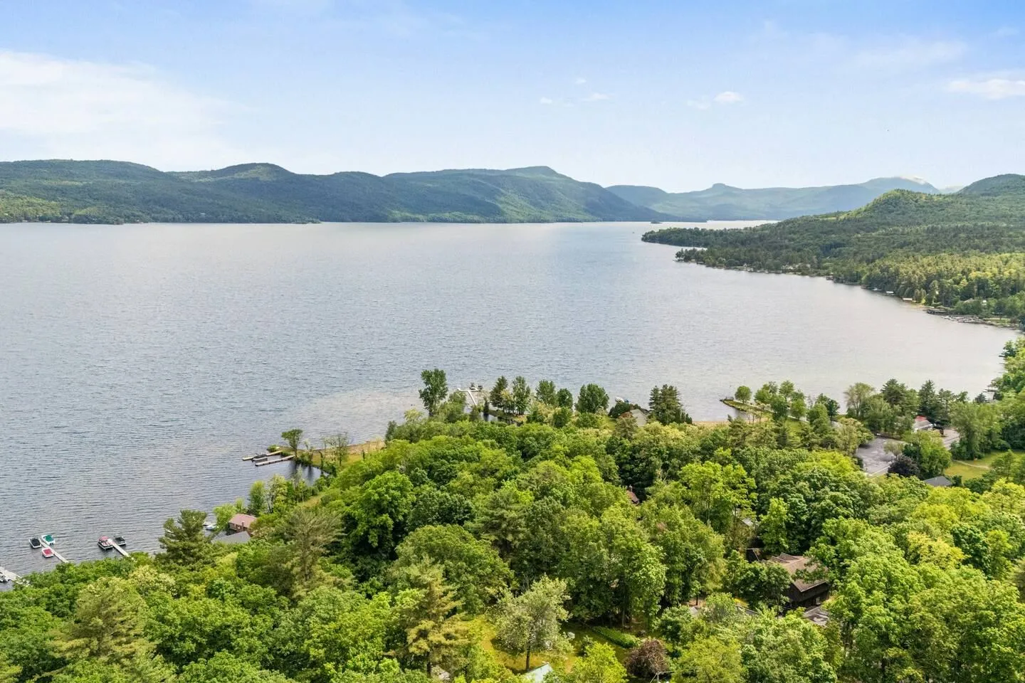 High aerial view overlooking a vast lake bordered by lush green trees, docks, and distant rolling mountains under a pale sky.