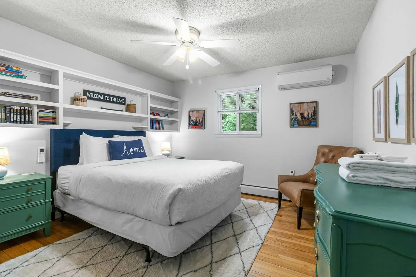 Bedroom featuring a made bed with a dark blue headboard beneath built-in shelving, a ceiling fan, hardwood floors, and a patterned rug.