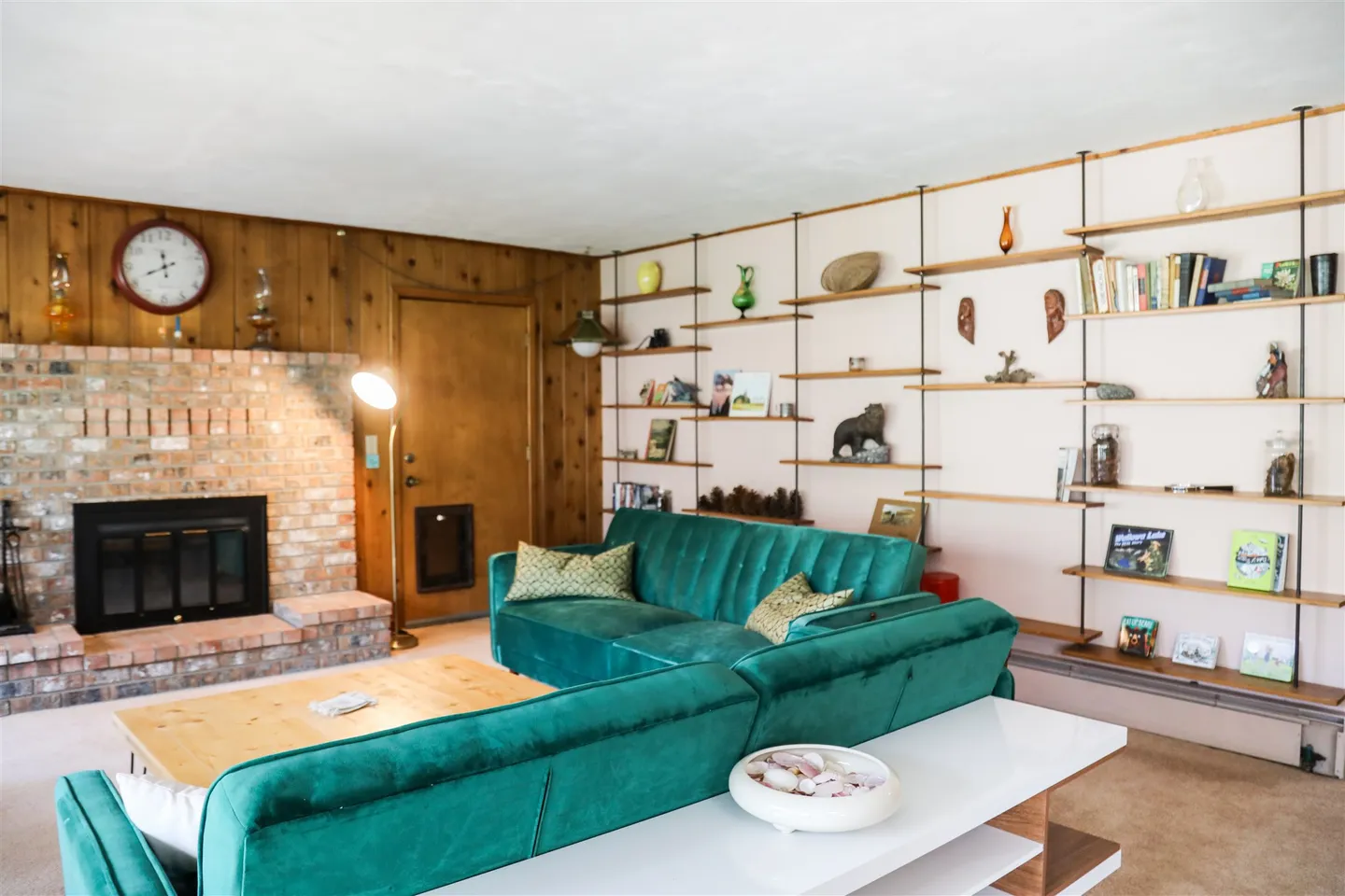 Living room with a green sectional couch, brick fireplace, wood-paneled wall, and wall-mounted display shelving.