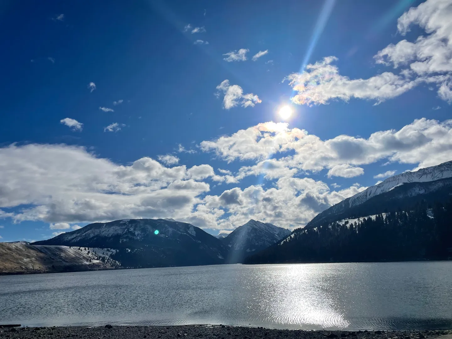 Sunlight reflecting off a wide lake surrounded by snow-dusted mountains under a blue sky with scattered clouds.