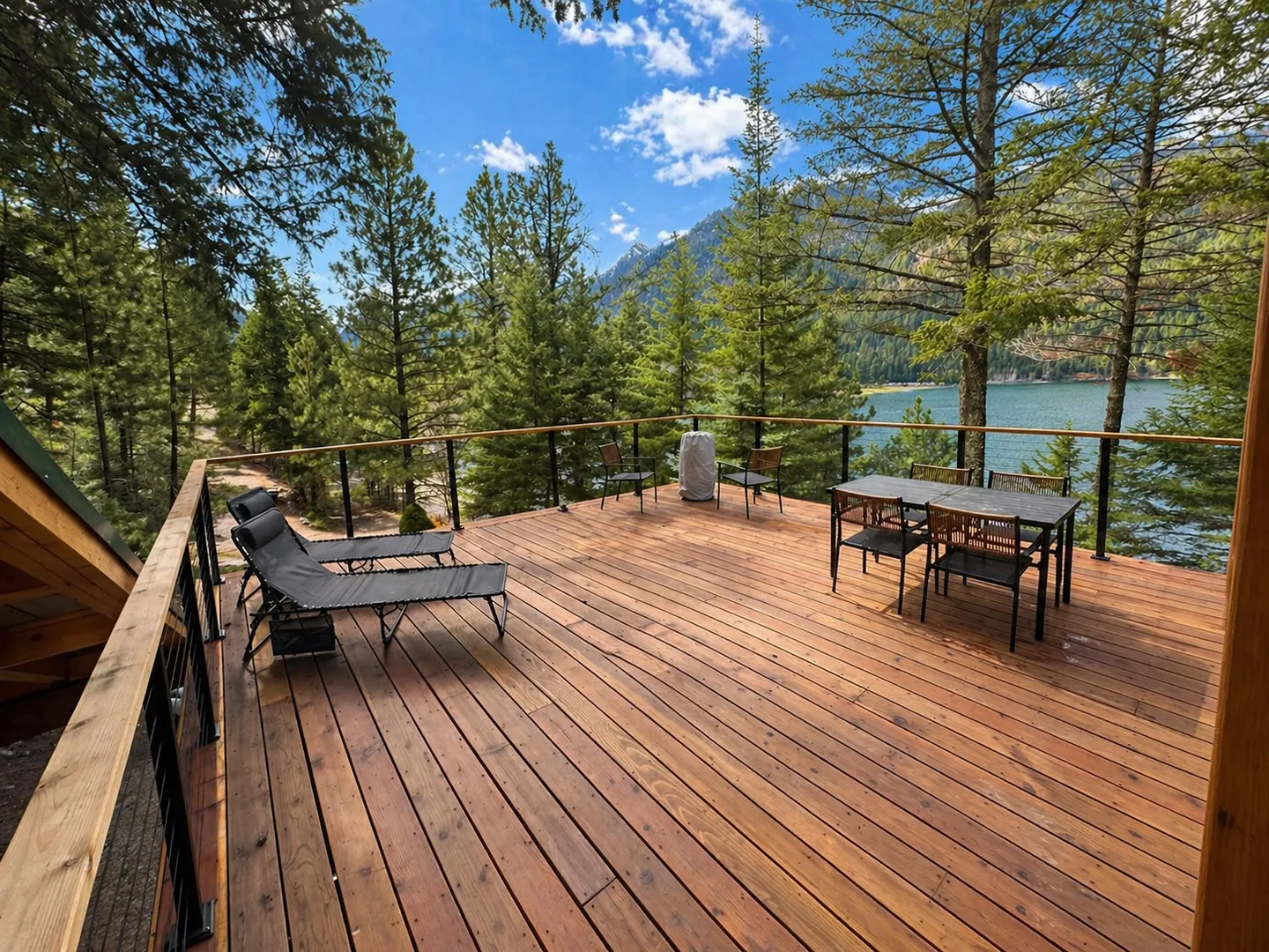 Spacious wood deck with lounge chairs, a dining set, and views of a lake and pine trees.