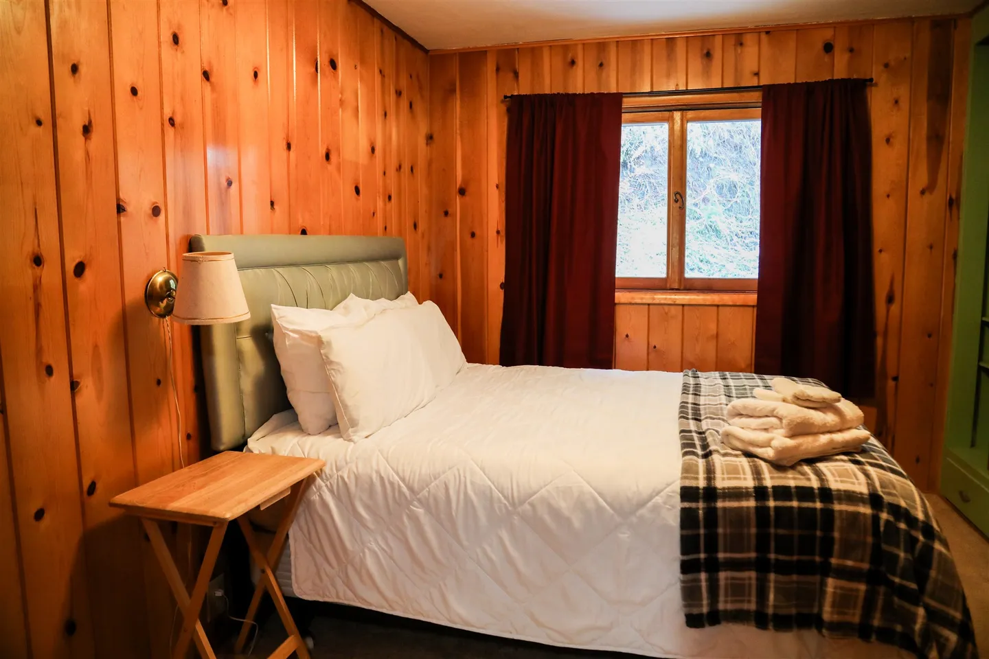 Bedroom with knotty wood-paneled walls, a made bed with white linens, and a window with dark red curtains.