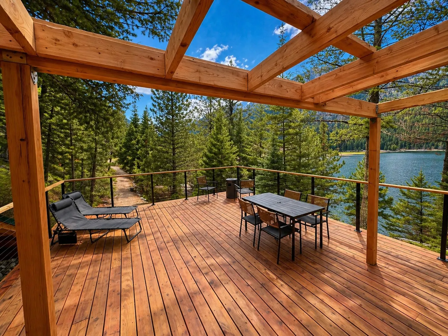 Spacious wooden outdoor deck under a timber pergola with a dining table, lounge chairs, and views of a lake and pine forest.