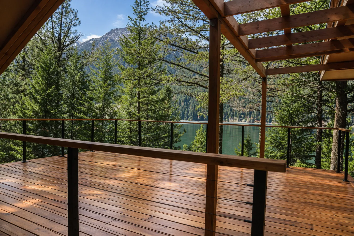 A wooden outdoor deck with modern railings overlooking evergreen pine trees, a calm lake, and a mountain peak in the distance.